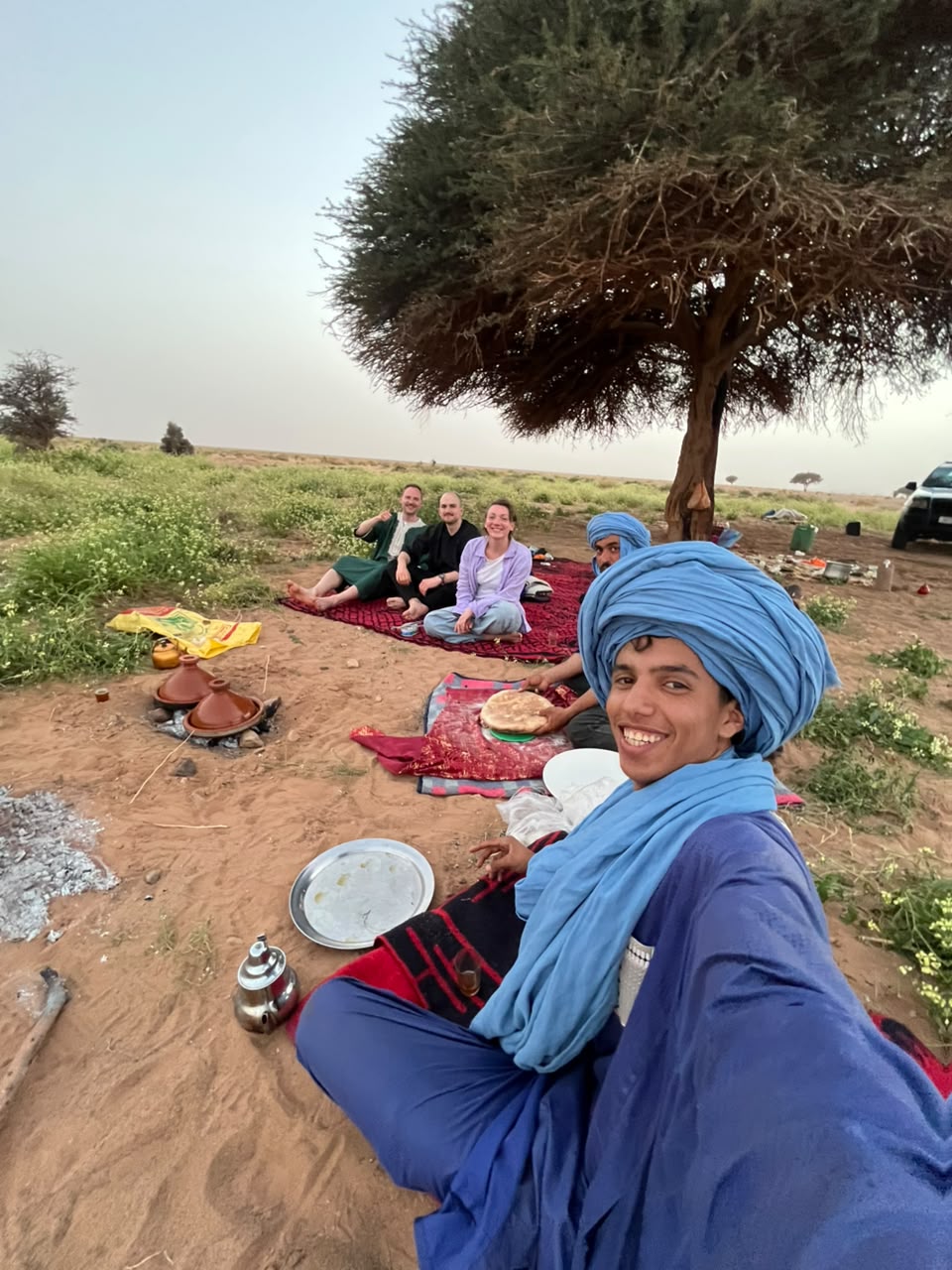 Our guide in a blue turban smiling, with travellers sharing a meal on rugs under an acacia